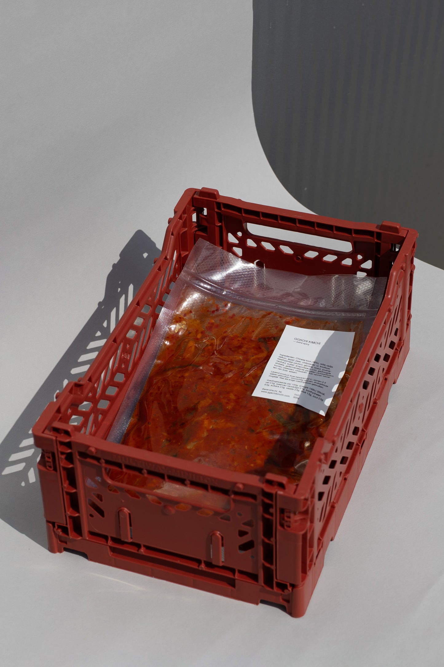 Red crate with a clear plastic bag containing bags of kimchi on a gray background. It's a kimchi tasting box consisting of 3 bags