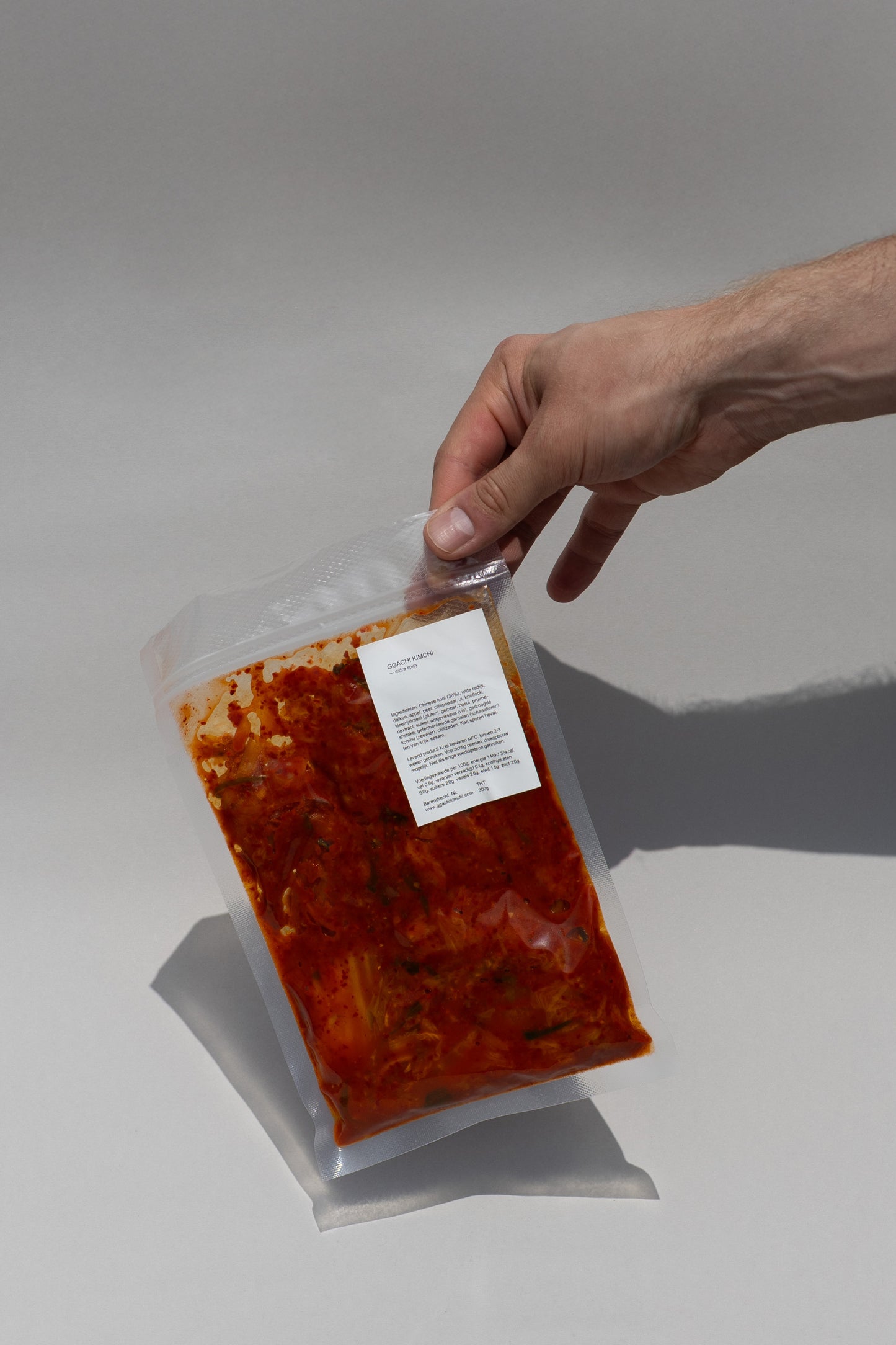 Hand holding a vacuum-sealed package of extra spicy kimchi with a label on a gray background. 